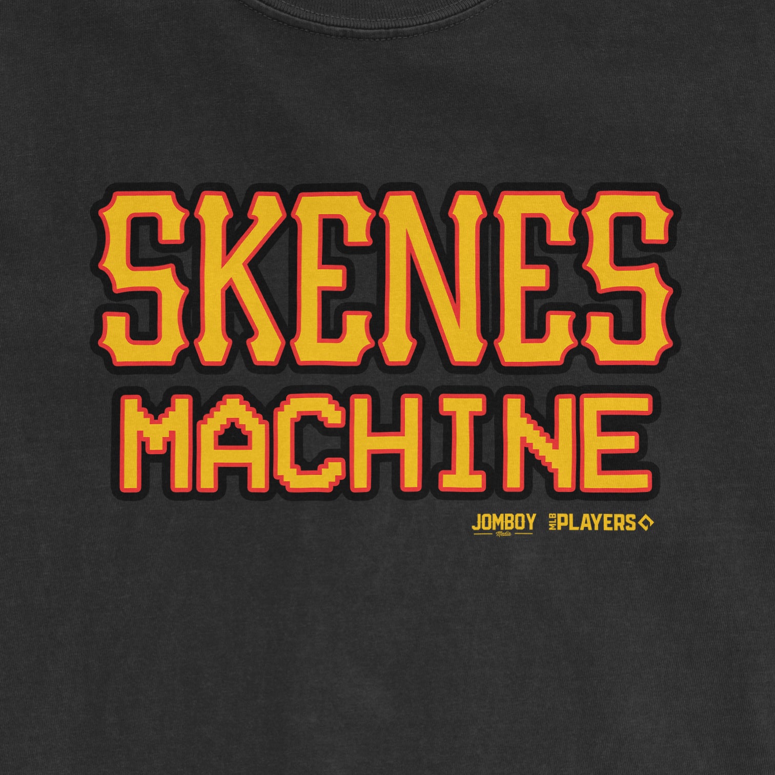 Close-up of a black T-shirt featuring bold yellow and red "SKENES MACHINE" text with smaller "JOMBOY" and "MLB PLAYERS" logos below.