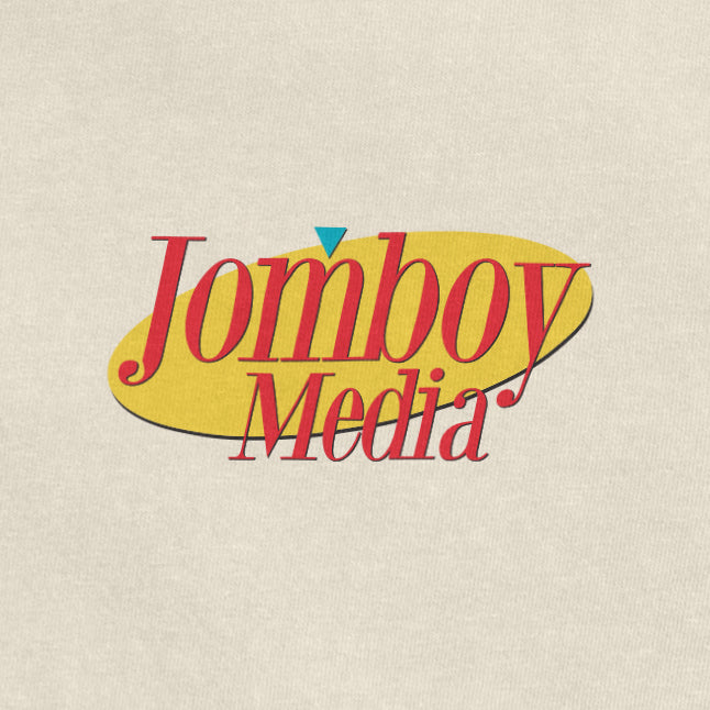 "Jomboy Media logo with red text, yellow oval background, and a small blue triangle above the 'J' on a light beige fabric.