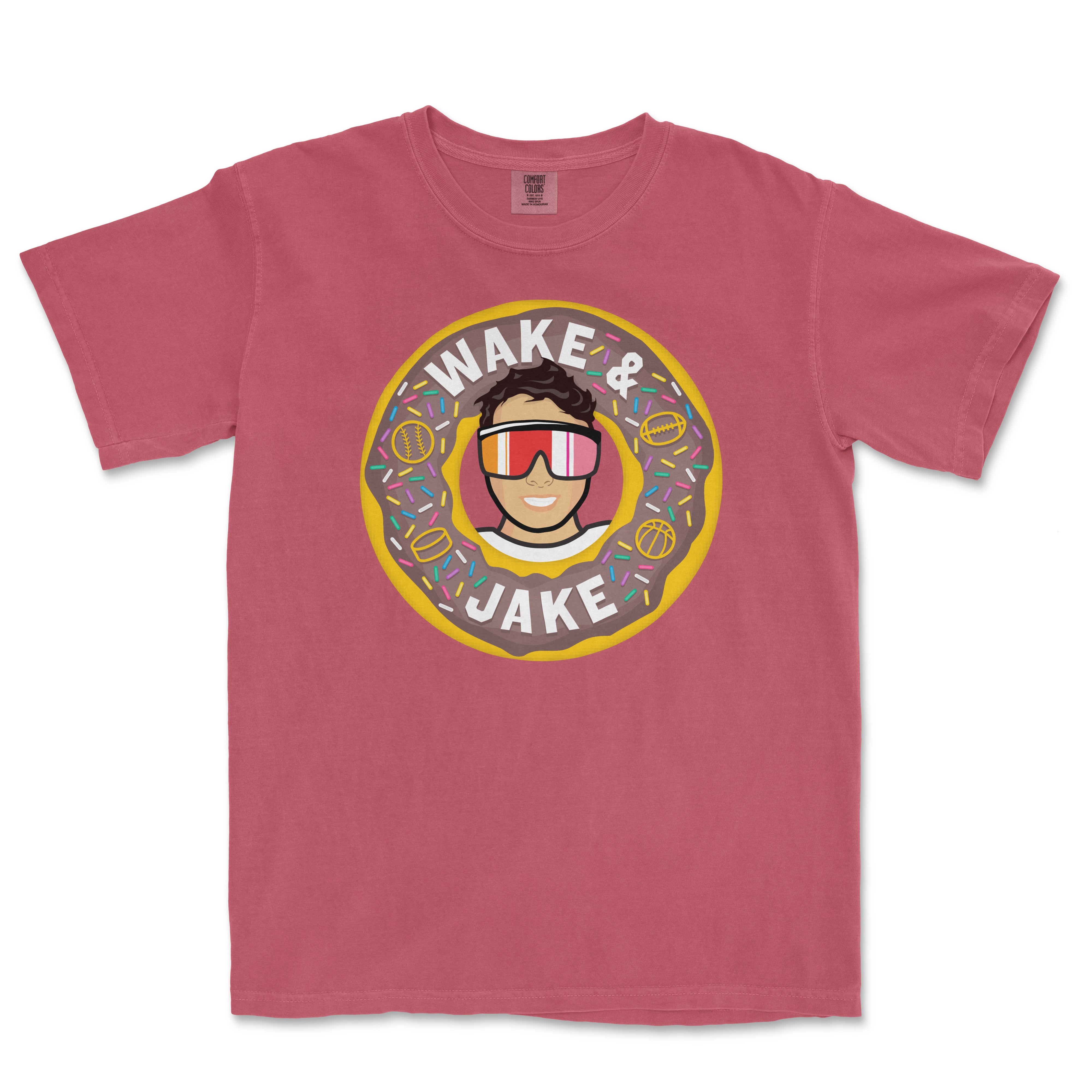 Pink T-shirt featuring a cartoon illustration of a smiling person wearing sunglasses, surrounded by a donut with sprinkles and sports icons, with "Wake & Jake" text.