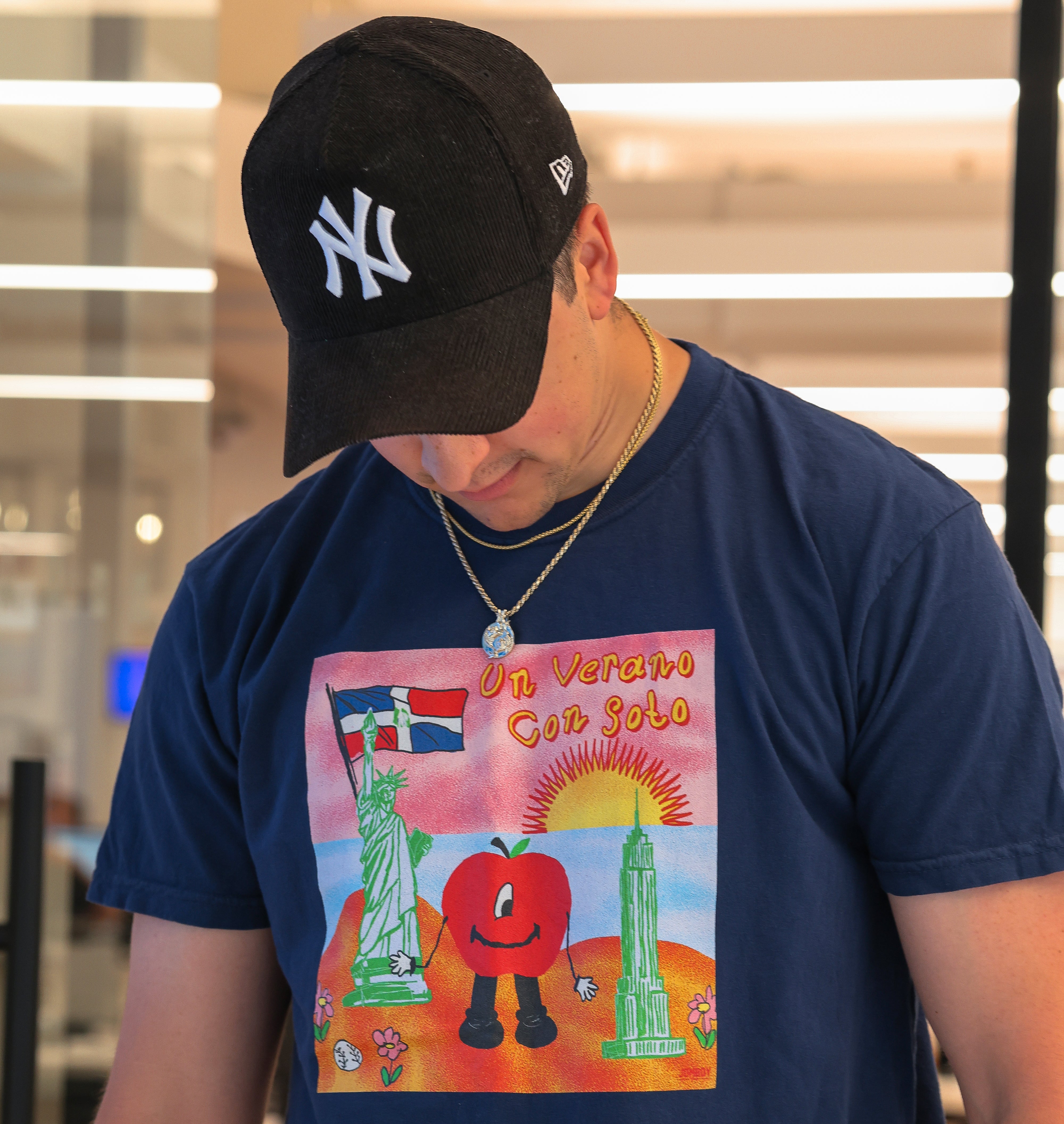Man wearing a black New York Yankees cap, navy T-shirt with colorful "Un Verano Con Soto" design, and layered necklaces.